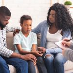 Bipolar Disorder In Children Concept. Little african american girl and her parents at reception of psychologist, kid sharing her feelings with doctor