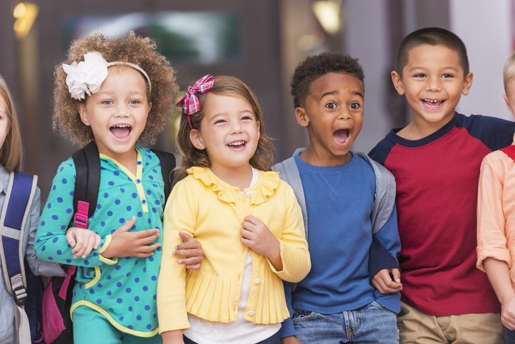 A multi-ethnic group of children standing in a row in a school hallway, excited and laughing, watching something. They are in kindergarten or preschool, carrying bookbags. They are 4 to 6 years old.
